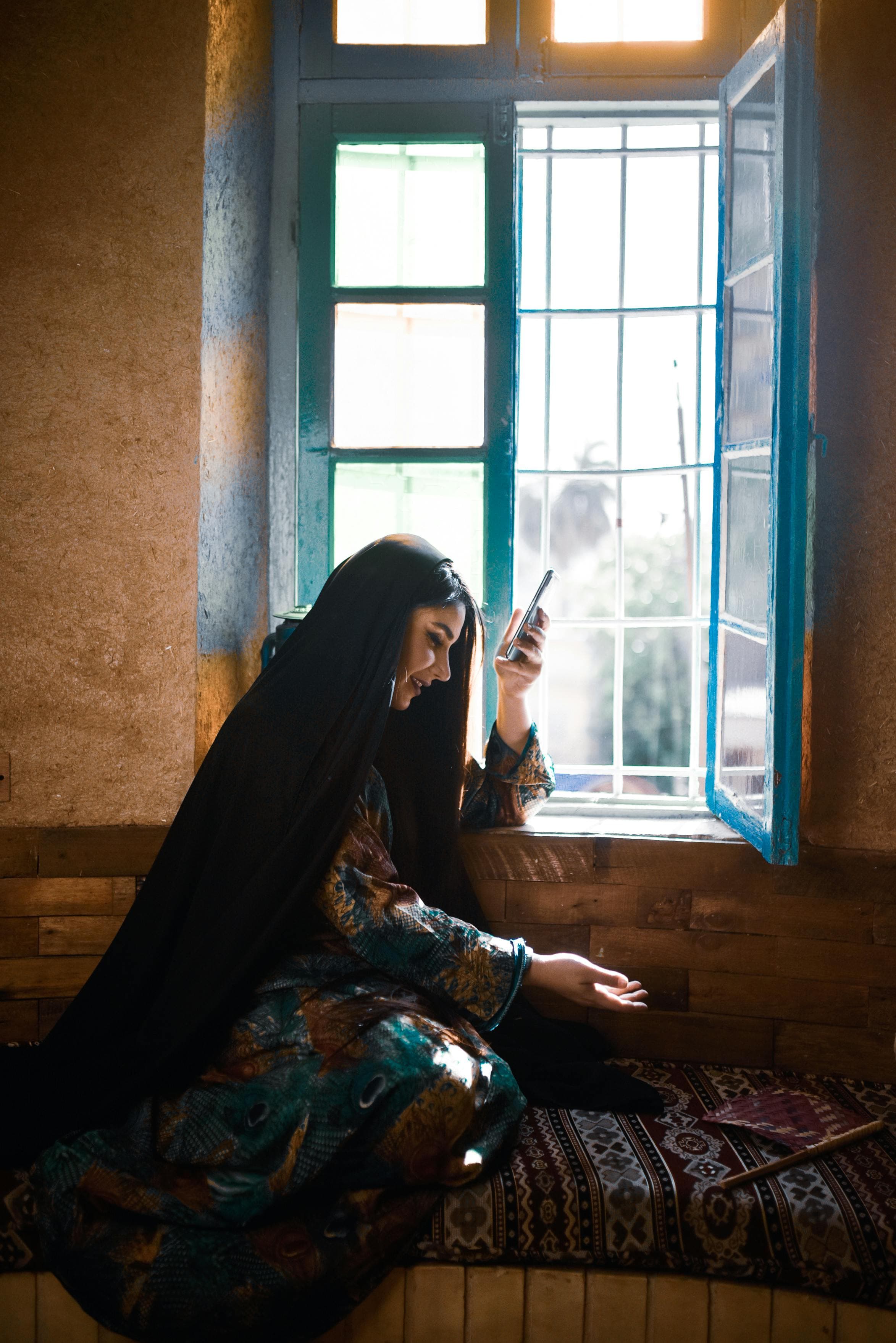Mulher afegã sentada perto de uma janela azul segurando o seu telefone