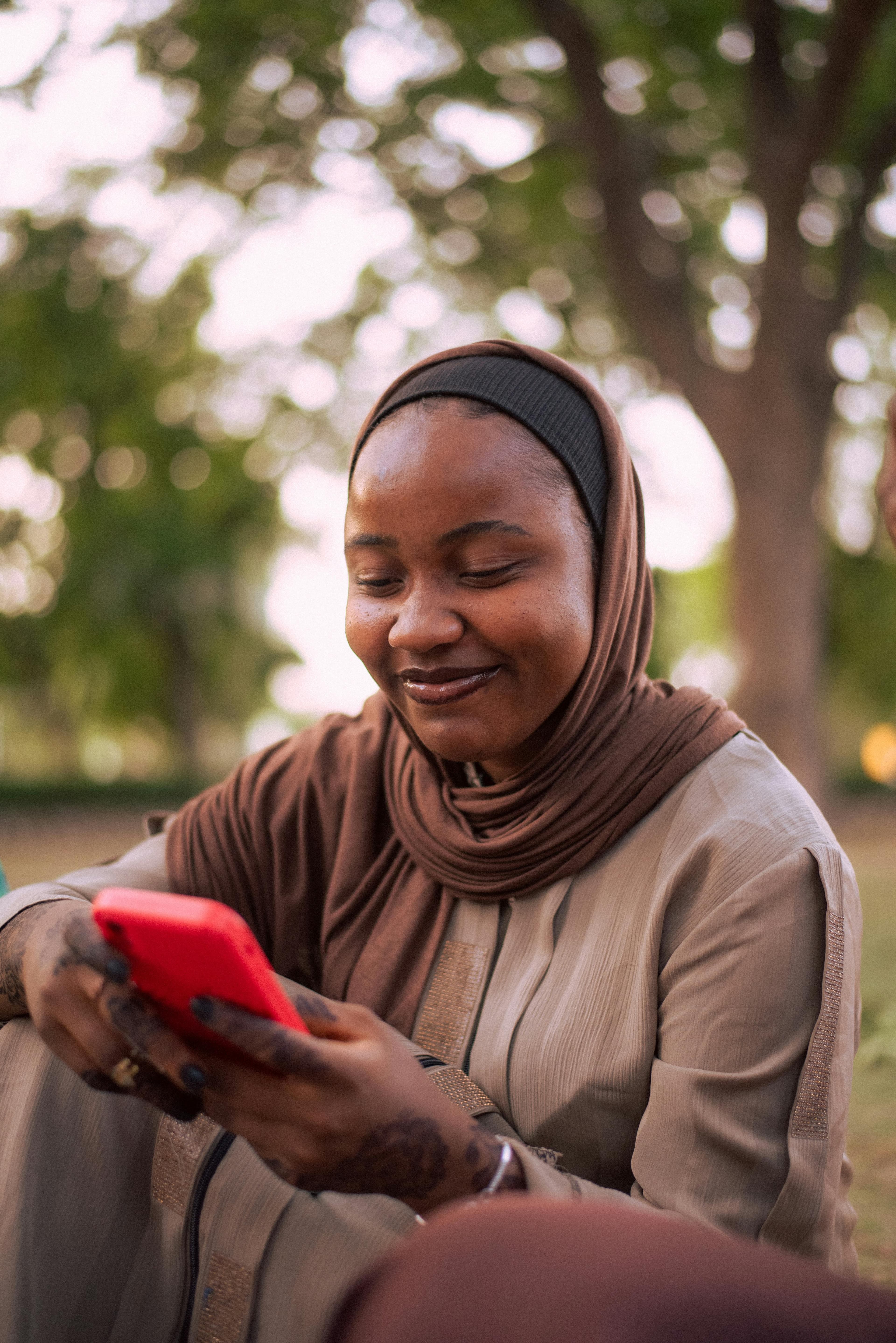 Donna con hijab marrone sorridente guarda il suo telefono rosso, esterni